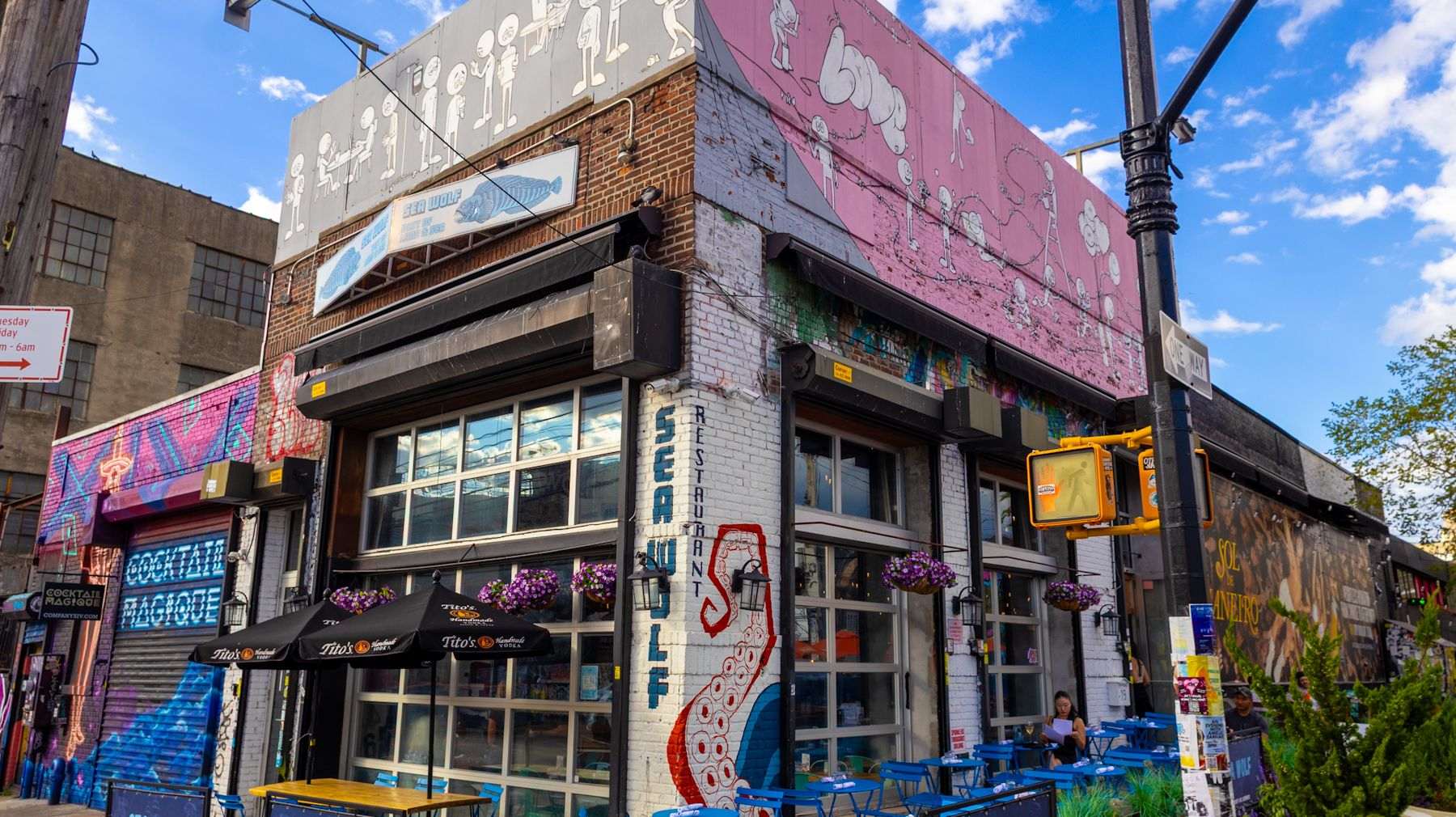 The Sea Wolf corner at Wyckoff Avenue with murals, blue sidewalk tables and hanging flowers